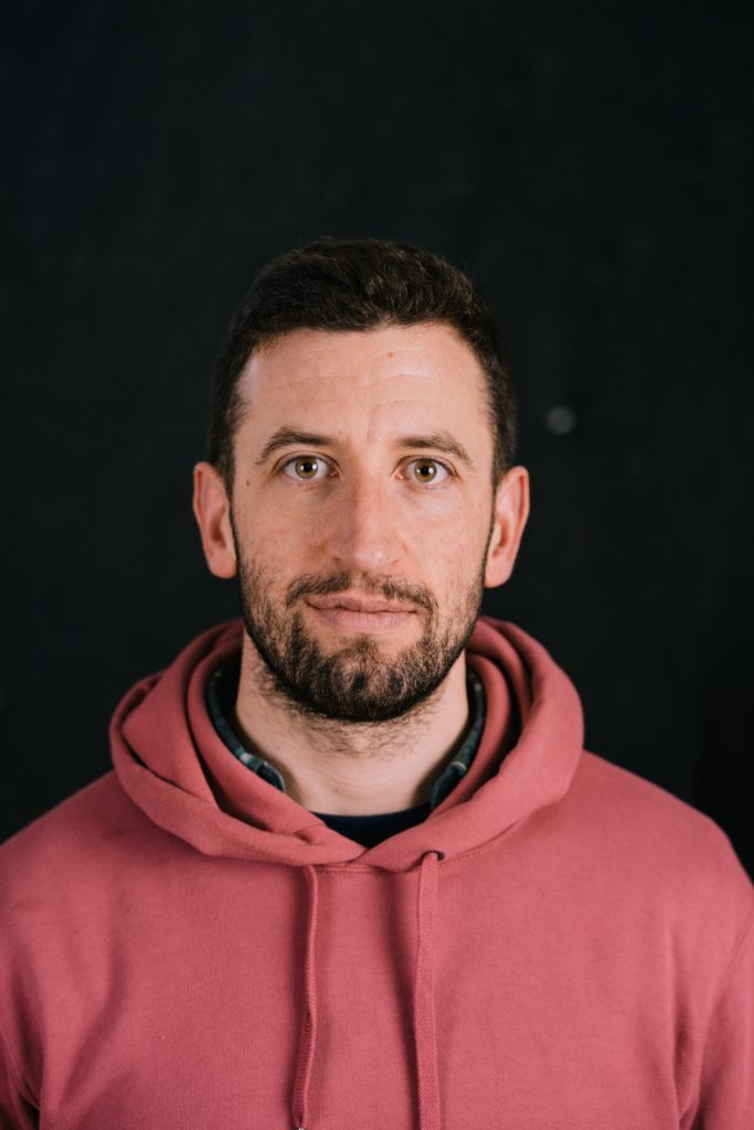 portrait image of Hugh wearing a terracotta hoodie smiling in front of a black background